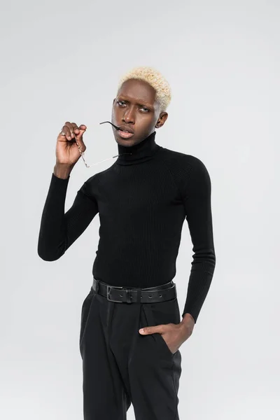 Blond afro-américain au col roulé tenant des lunettes de soleil isolées sur gris — Photo de stock