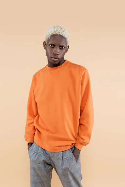 Blonde african american man in orange sweatshirt posing with hands in pockets isolated on beige — Stock Photo