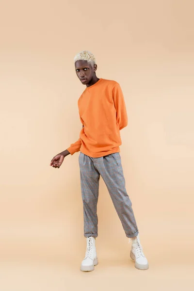 Piena lunghezza di uomo africano americano biondo in felpa arancione in piedi e in posa sul beige — Foto stock