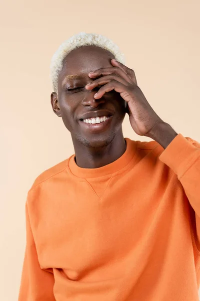 Fröhlicher afrikanisch-amerikanischer Mann in orangefarbenem Sweatshirt, lächelnd und mit verdeckenden Augen isoliert auf beige — Stockfoto