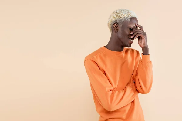 Shy african american man in orange sweatshirt smiling and covering eyes isolated on beige — Stock Photo