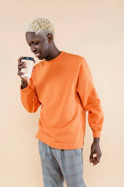 Американский блондин в оранжевой толстовке улыбается, держа в руках бумажную чашку, изолированную на бежевом — стоковое фото