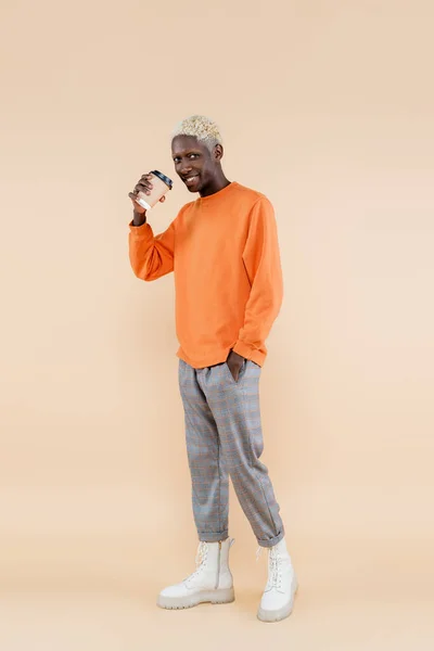 Full length of blonde african american man in orange sweatshirt smiling while holding paper cup and posing on beige — Stock Photo