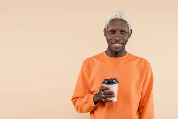 Hombre afroamericano feliz en sudadera naranja sonriendo mientras sostiene taza de papel aislado en beige - foto de stock