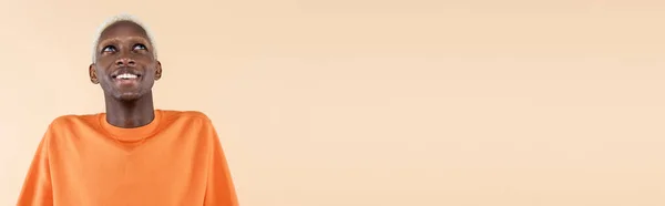 Счастливый африканский американец в оранжевой толстовке улыбается, глядя на изолированный бежевый баннер — стоковое фото