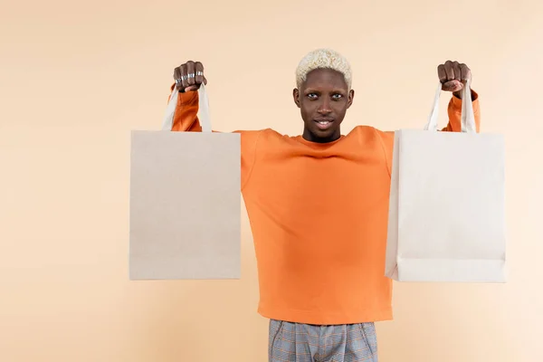 Smiling african american man in orange sweatshirt holding shopping bags isolated on beige — Stock Photo