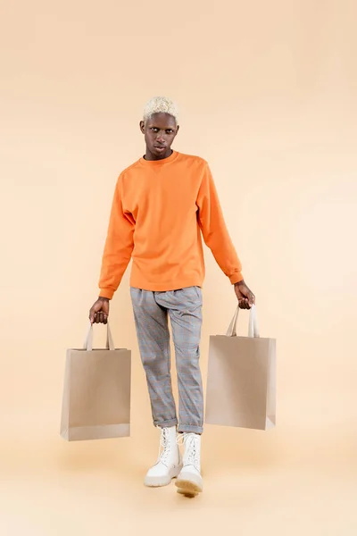 Full length of young african american man in orange sweatshirt holding shopping bags on beige — Stock Photo