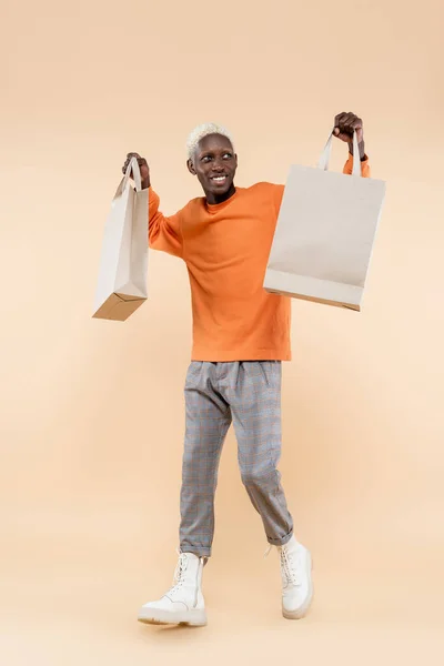 Full length of happy african american man in orange sweatshirt holding shopping bags on beige — Stock Photo
