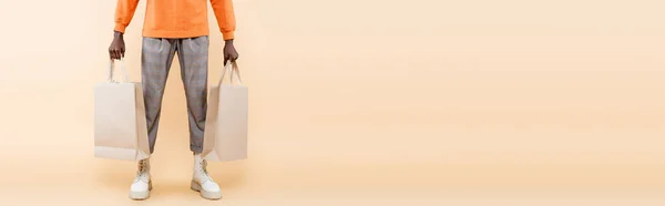 Cropped view of african american man in orange sweatshirt holding shopping bags on beige, banner — Stock Photo