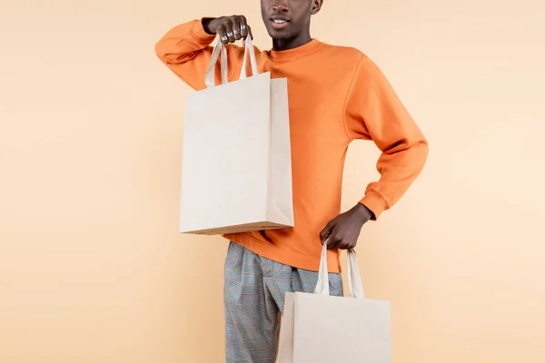 Обрізаний вид на молодого афроамериканця в помаранчевому светрі, що тримає сумки для покупок ізольовані на бежевому — стокове фото
