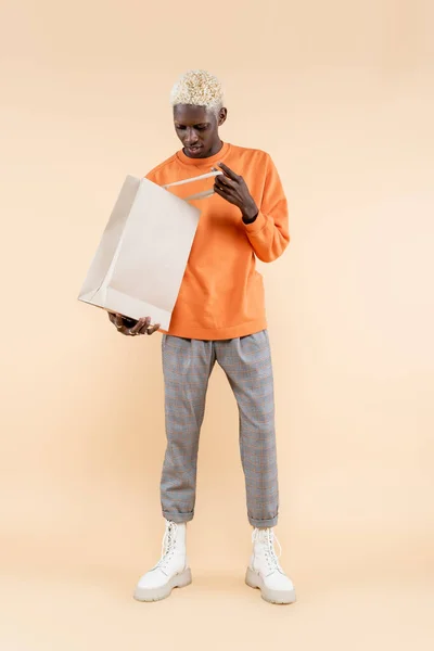 Full length of young african american man in orange sweatshirt holding shopping bag on beige — Stock Photo