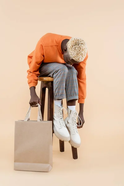 Comprimento total de homem americano africano loiro em camisola laranja segurando saco de compras e sentado na cadeira no bege — Fotografia de Stock