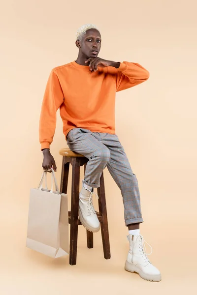 Voller Länge junger afrikanisch-amerikanischer Mann in orangefarbenem Sweatshirt mit Einkaufstasche und auf einem Stuhl auf beige sitzend — Stockfoto