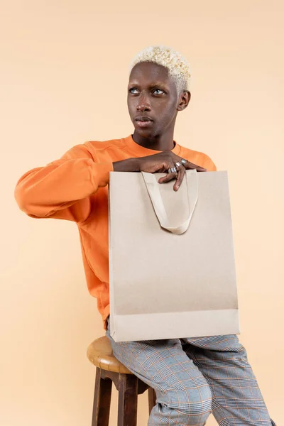 Loira homem americano africano em camisola laranja segurando saco de compras e sentado na cadeira isolada no bege — Fotografia de Stock