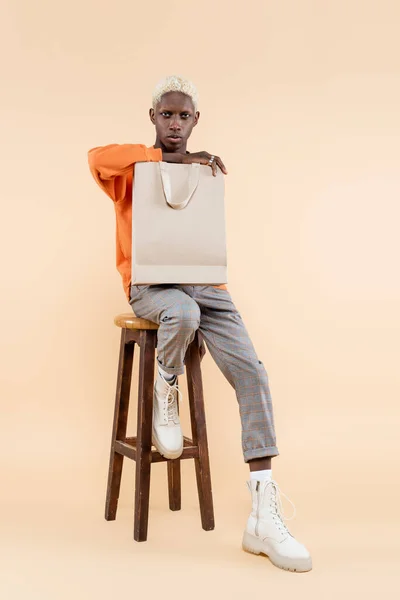 Full length of blonde african american man in sweatshirt holding shopping bag and sitting on chair on beige — Stock Photo