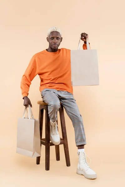 Full length of blonde african american man in sweatshirt holding shopping bags and sitting on chair on beige — Stock Photo