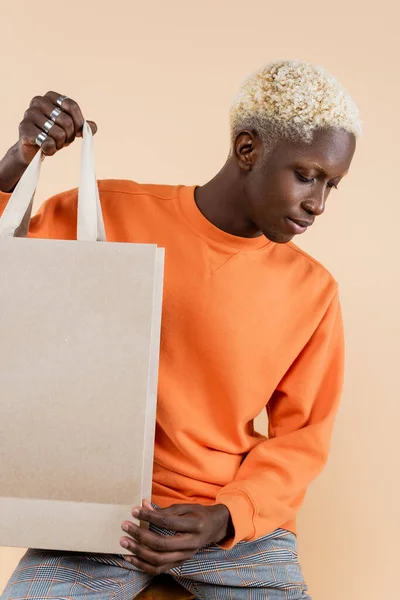 Blonde african american man in sweatshirt holding shopping bag isolated on beige — Stock Photo
