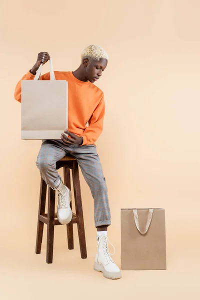 Full length of blonde african american man in sweatshirt looking at shopping bag and sitting on chair on beige — Stock Photo