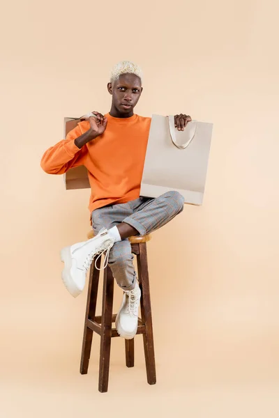 Full length of young african american man in sweatshirt sitting on chair with shopping bags on beige — Stock Photo