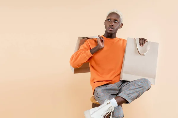 Joven afroamericano hombre en sudadera sentado en silla con bolsas aisladas en beige - foto de stock
