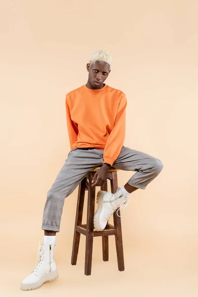 In voller Länge junger afrikanisch-amerikanischer Mann im Sweatshirt sitzt auf einem Stuhl auf beige — Stockfoto