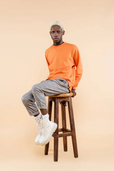 Повна довжина блондинки афроамериканця в светрі, сидячи на стільці на бежевому — стокове фото