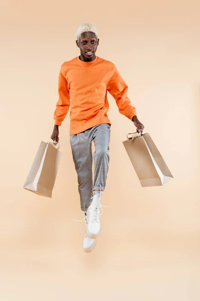 Longitud completa de hombre afroamericano rubio en sudadera levitando y sosteniendo bolsas de compras en beige - foto de stock