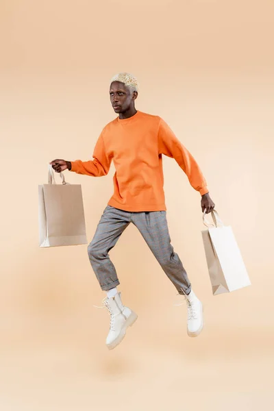 Full length of blonde african american man in sweatshirt levitating while holding shopping bags on beige — Stock Photo