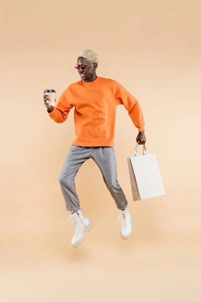 Pleine longueur de heureux homme afro-américain en lunettes de soleil lévitant tout en tenant tasse en papier et sac à provisions sur beige — Photo de stock