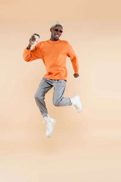Longitud completa de hombre afroamericano feliz en gafas de sol levitando mientras sostiene la taza de papel en beige - foto de stock