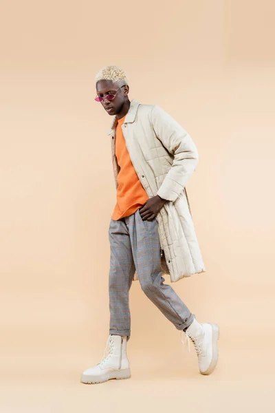 Full length of young african american man in stylish sunglasses and coat posing with hand in pocket on beige — Stock Photo