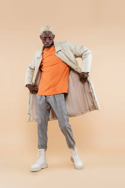 Повна довжина стильного афроамериканця в сонцезахисних окулярах і пальто позує на бежевому — стокове фото