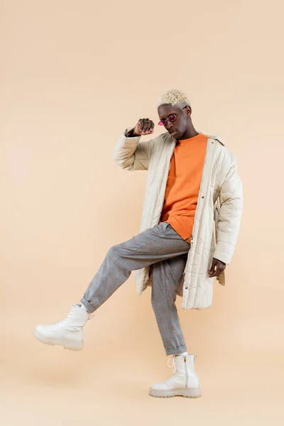 Full length of young african american man in trendy sunglasses and coat posing on beige — Stock Photo