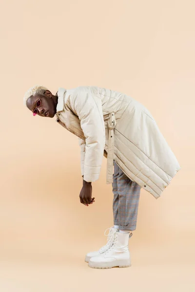 Full length of young african american man in stylish sunglasses and coat bending while posing on beige — Stock Photo