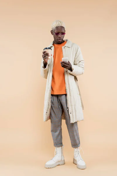 Pleine longueur de jeune homme afro-américain dans des lunettes de soleil élégantes et manteau à l'aide d'un smartphone et tenant tasse en papier sur beige — Photo de stock