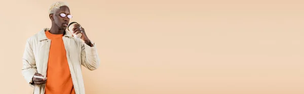 Jeune homme afro-américain dans des lunettes de soleil élégantes et manteau tenant smartphone et tasse en papier isolé sur beige, bannière — Photo de stock
