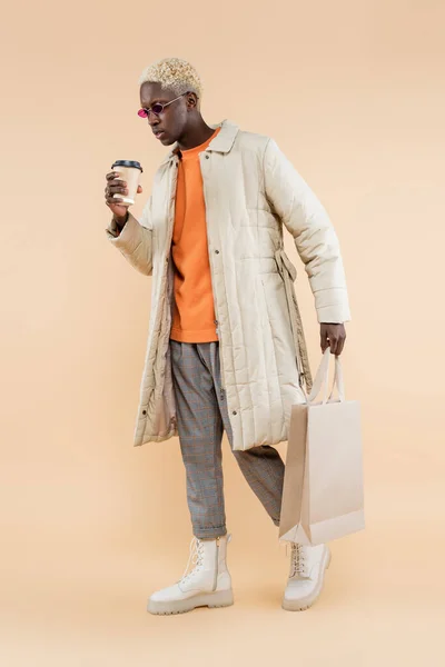 Full length of young african american man in coat holding paper cup and shopping bag on beige — Stock Photo