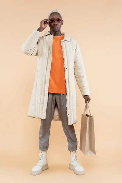 Full length of young african american man in coat holding shopping bag while adjusting sunglasses on beige — Stock Photo