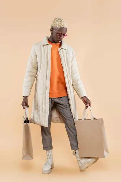 Full length of young african american man in coat and sunglasses holding shopping bags on beige — Stock Photo