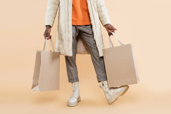 Vista recortada de hombre afroamericano en abrigo sosteniendo bolsas de compras en beige - foto de stock