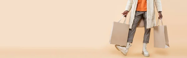 Cropped view of african american man in coat holding shopping bags on beige, banner — Stock Photo