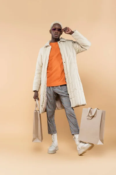 Full length of trendy african american man in coat adjusting sunglasses and holding shopping bag on beige — Stock Photo