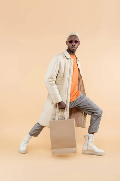 Longitud completa de hombre afroamericano de moda en abrigo y gafas de sol posando con bolsa de compras en beige - foto de stock