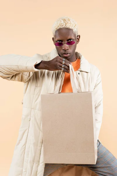 Блондинка афроамериканка в стильному пальто і модні сонцезахисні окуляри тримає сумку для покупок ізольовано на бежевому — стокове фото