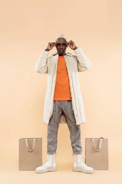 Full length of trendy african american man in stylish coat adjusting sunglasses near shopping bags on beige — Stock Photo