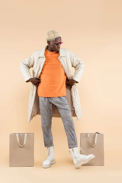 Full length of trendy african american man in stylish coat and sunglasses posing near shopping bags on beige — Stock Photo
