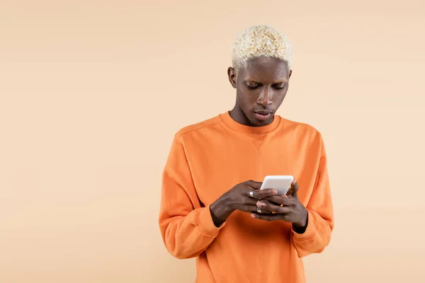 Blonder afrikanisch-amerikanischer Mann in orangefarbenem Sweatshirt mit Smartphone isoliert auf beige — Stockfoto