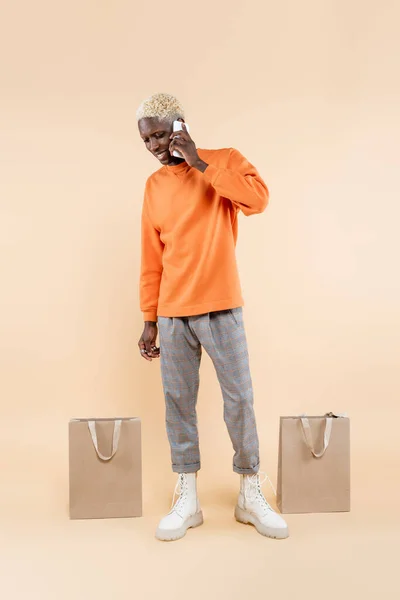 Longitud completa de hombre afroamericano feliz en sudadera naranja hablando en el teléfono inteligente cerca de bolsas de compras en beige - foto de stock
