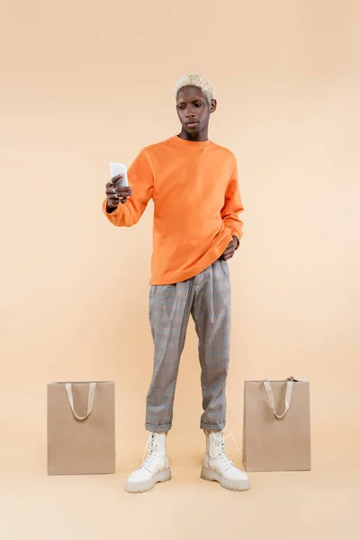 Comprimento total do homem americano africano em camisola laranja levando selfie no smartphone perto de sacos de compras em bege — Fotografia de Stock