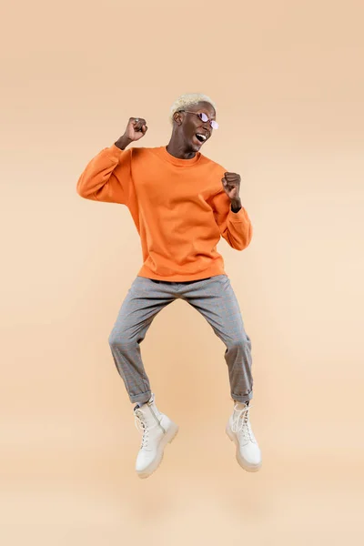 Full length of excited african american man in orange sweatshirt and sunglasses jumping on beige — Stock Photo